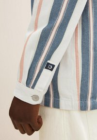 Jasnoniebieska denimowa kurtka w poziome paski w kolorach niebieskim, różowym i białym. Posiada zapięcie na guzik przy mankietach oraz małą metkę z logo na rękawie.