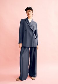 Giacca doppiopetto in carbone abbinata a pantaloni a gamba larga; tessuto liscio, vestibilità oversize, completata da un berretto da baseball nero. Sfondo rosa.