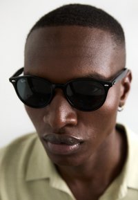 Ochelari de soare negri cu un finisaj lucios și lentile rotunde. Au un pod proeminent și un logo Ray-Ban subtil, purtați cu un tricou polo verde deschis.