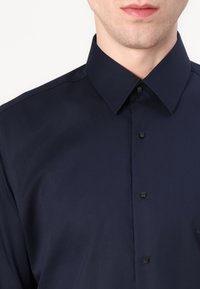 Sötétkék, klasszikus galléros ing, fekete négyzet alakú gombokkal és sima textúrával, amely hangsúlyozza a testre szabott illeszkedést.