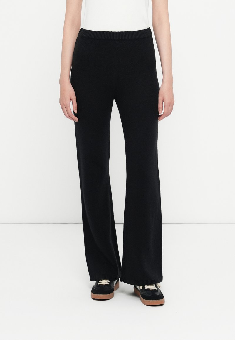 Pantalons larges noirs en tissu tricoté doux, avec une ceinture côtelée et une texture lisse, associés à des chaussures noires et un haut blanc.