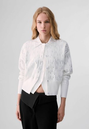 Młoda kobieta o blond włosach, ubrana w białą, zapinaną na guziki kurtkę z srebrnymi ozdobami oraz czarne spodnie z wysokim stanem na jednolitym tle.