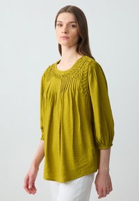 Blusa verde oliva con maniche a tre quarti, con collo ricamato e dettagli plissettati. Realizzata in tessuto texturizzato, drappeggiata delicatamente.