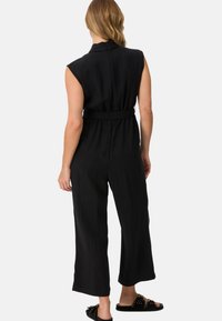 Svart ärmlös jumpsuit med inbakad midja, vid passform och släthet i texturen. Baksidan har en hög halsringning och minimalt med detaljer.