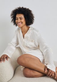 Camicia di colore crema, dal taglio morbido con tessuto testurizzato e motivi sottili, maniche lunghe e dettagli con tasca. Indossata con le gambe incrociate su una superficie morbida.