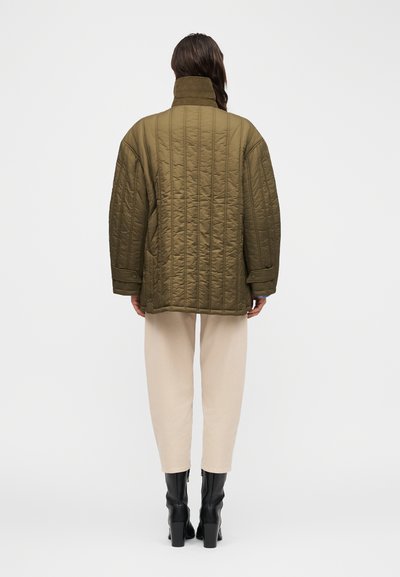 Άτομο με υπερμεγέθη πράσινο χακί μπουφάν με καπιτονέ σχέδιο, μπεζ παντελόνι και μαύρες μπότες με τακούνι, στέκεται πλάτη προς την κάμερα.