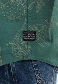 Chemise verte avec imprimé floral beige et étiquette noire « Petrol Ind & Co. » cousue près de l’ourlet sur un jean denim bleu.