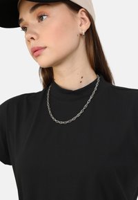T-shirt nera con un collo alto, abbinata a una collana di argento. Il modello indossa un cappellino e orecchini a cerchio, con capelli e pelle lisci.