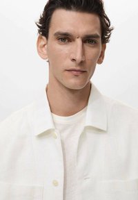 White, textured shirt with a button-up collar, worn over a light gray T-shirt; features a single chest pocket and subtle stripe pattern.