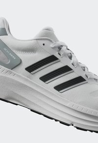 Zapatilla deportiva blanca con un upper texturizado, tres franjas negras y plateadas, y una suela gruesa. Presenta un acento gris en el talón.