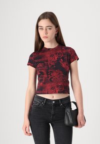 Camiseta roja de culto con texto y diseños gráficos en negro, manga corta, combinada con jeans negros de talle alto y un pequeño bolso negro.