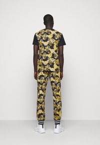 Mann in schwarz-weißen und gelb gemusterten Hosen sowie passendem ärmellosem Oberteil über einem schwarzen T-Shirt, der mit dem Rücken zur Kamera auf einem weißen Hintergrund steht.