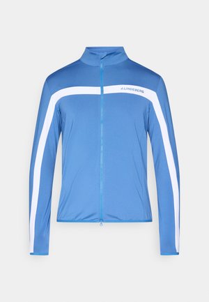 Blå atletisk jakke lavet af glat stof, med hvide diagonale striber, en høj krave og en fuld lynlås foran.