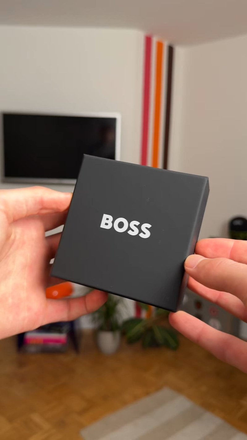 Черна кутия с гладко покритие и надпис "BOSS" в удебелен бял шрифт. Държана между две ръце, с текстуриран фон.