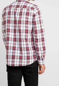 Uomo che indossa una camicia a maniche lunghe bianca, rossa e nera a scacchi con pantaloni neri, visto da dietro su uno sfondo neutro.