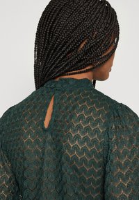 Grön spetsblus med mönstrad textur, nyckelhål baktill och korta puffärmar. Materialet verkar lätt och genomskinligt.