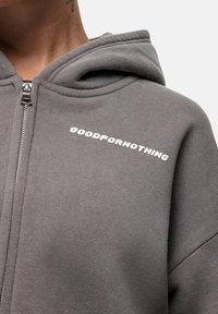 Felpa grigia con zip realizzata in tessuto morbido, dotata di cappuccio e testo bianco "GOODFORNOTHING" nella zona sinistra del petto.
