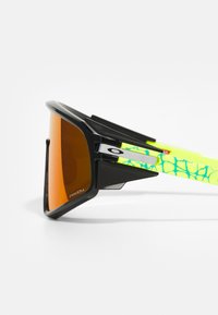 Óculos de sol pretos com lentes laranja, design angular e ousado, hastes com padrão verde e azul, e marcação proeminente na armação.