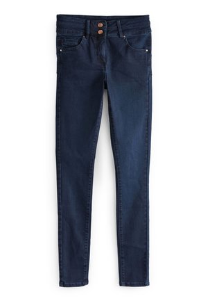 Femme portant un haut asymétrique vert olive, un jean skinny foncé avec fermeture à double bouton et des sandales à talons hauts dorées devant un mur rose.