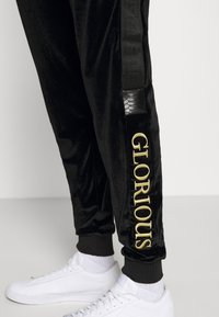 Schwarze Velour-Jogginghosen mit "GLORIOUS" in Gold auf dem Bein, kombiniert mit weißen Sneakers und sichtbaren Rippbündchen. Minimalistischer Hintergrund.