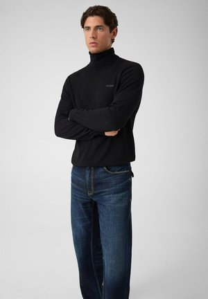 Uomo con le braccia incrociate che indossa un maglione a collo alto nero Guess e jeans blu, in piedi davanti a uno sfondo semplice grigio chiaro.