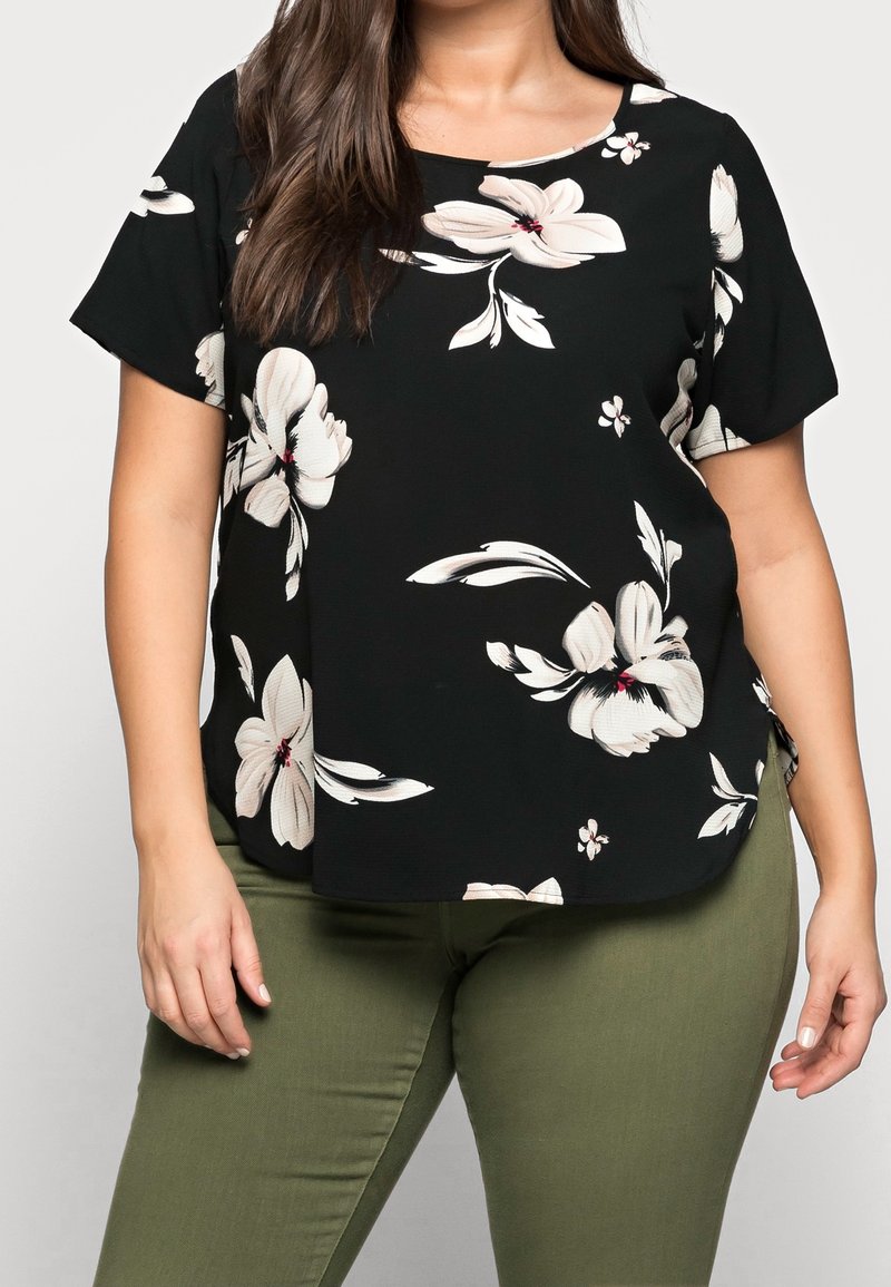 Femme portant un haut noir à manches courtes avec de grandes impressions florales blanches associé à un pantalon ajusté vert olive, visible du cou aux hanches.