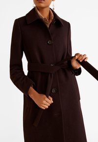 Hnědý vlněný kabát s přiléhavým střihem, který má límec, zapínání na knoflíky a stahovací pásek, doplněný o boční kapsy a hladkou texturu.