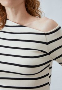 Lange ermetopp i krem med svarte horisontale striper, med en asymmetrisk off-the-shoulder halslinje og myk, stretchy stoff.