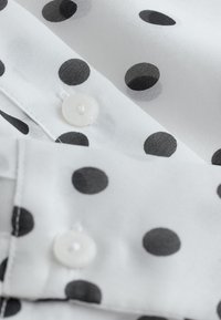 Tissu blanc à pois noirs montrant deux boutons blancs cousus et des bords surpiqués, suggérant une chemise ou un chemisier à motif.