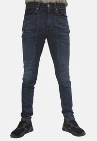 Jeans in denim blu scuro con vestibilità slim, caratterizzati da tasche frontali distintive e una texture liscia, abbinati a scarpe sportive nere.