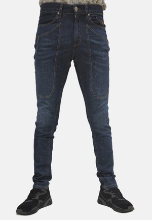 Jeans in denim blu scuro con vestibilità slim, caratterizzati da tasche frontali distintive e una texture liscia, abbinati a scarpe sportive nere.