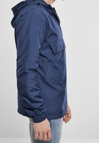 Granatowa kurtka z kapturem z zamkiem przy szyi, elastycznymi mankietami i kieszenią na przodzie. Gładka faktura i lekki materiał.