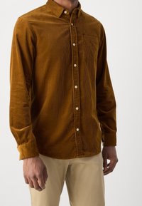 Camisa marrón de pana con cuello abotonado, bolsillo delantero y botones beige. Presenta una textura acanalada y mangas largas.