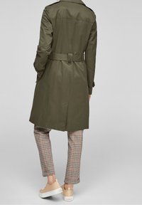 Trench coat verde oliva con vita con cintura, polsini abbottonati e schiena ventilata. Indossato sopra pantaloni a quadri e sneaker beige.