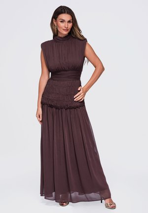 Mujer con cabello largo lleva un vestido maxi plisado sin mangas de color marrón oscuro y tacones dorados abiertos en los dedos, de pie con una mano en la cadera.