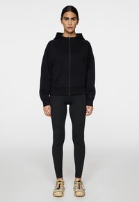 Schwarze Zip-Hoodie mit Kapuze, kombiniert mit schwarzen Leggings und beigen Sneakern. Glatte Textur und figurbetontes Design, mit kontrastierenden Details.