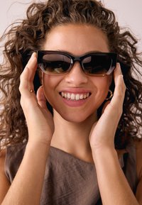 Schwarze Cat-Eye-Sonnenbrille mit Verlaufsgläsern und glänzendem Rahmen, getragen, während die Hände die Seiten halten. Das Modell hat lockiges Haar und trägt braune Kleidung.