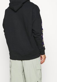 Felpa nera con cappuccio, caratterizzata da motivi geometrici colorati lungo le maniche. Tessuto morbido e polsini a costine. Abbinata a pantaloni cargo chiari.