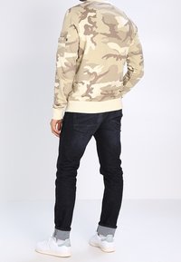 Svetří s crew neck v tan maskáčovém vzoru, tmavě modré džíny, bílé tenisky s šedými akcenty, viditelné švy a žebrované manžety.