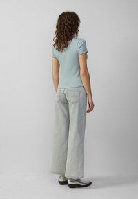 Frau mit lockigem Haar, die ein hellblaues Kurzarmoberteil, ausgebleichte weite Jeans und silberne Stiefeletten trägt, steht vor einer schlichten Wand.