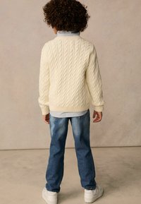 Cremefarbener Strickpullover mit Kabelmuster und gerippten Bündchen, getragen über einem hell gestreiften Hemd, kombiniert mit ausgebleichten blauen Jeans und weißen Sneakern.