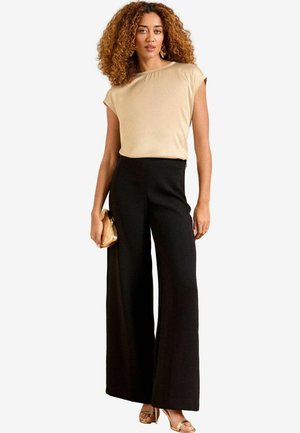 Beige kortom mouwtop gecombineerd met hoge zwarte wijde broek. Het model houdt een gouden clutch vast, wat de gladde stof textuur laat zien.
