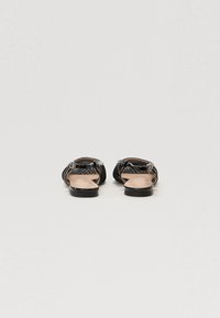 Sandales plates noires avec un design en maille, dotées d'une fine bride de cheville et d'une semelle lisse. L'intérieur est beige avec une texture subtile.
