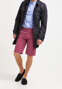 Black trench coat over a light blue shirt, paired with pink shorts. Black shoes with laces. Smooth textures and contrasting colors.
