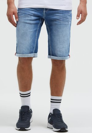 Man wearing rolled-up light blue denim shorts, white socks with black stripes, and navy blue sneakers, standing against a plain background.