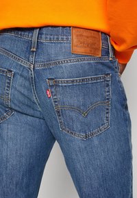 Blå denimjeans med en läderpatch i midjan, med två bakfickor och gula sömmar. Tyget har en klassisk texturerad finish.