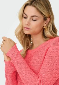 Femme blonde portant des boucles d'oreilles en or et un pull corail texturé, regardant vers le bas tout en ajustant la manche de son bras gauche.