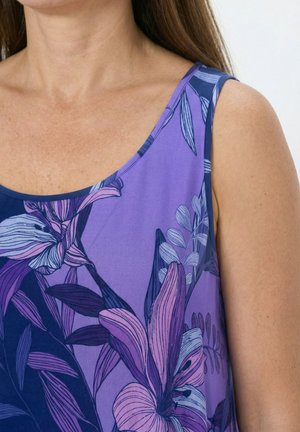 Donna che indossa una canotta viola senza maniche con motivi floreali blu e rosa, che mostra la spalla e la parte superiore del petto su uno sfondo chiaro.