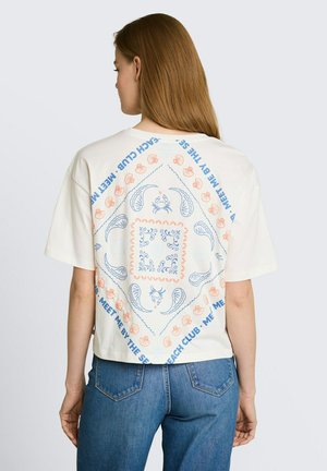 Vrouw met lang bruin haar die een wit T-shirt draagt met blauw en oranje zee-thema patronen en tekst, gecombineerd met een blauwe spijkerbroek, gezien van achteren.