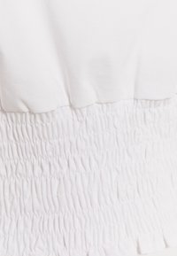 Top in tessuto bianco con vita arricciata e smockata, caratterizzata da una superficie testurizzata. Il materiale è liscio e presenta un colore uniforme.